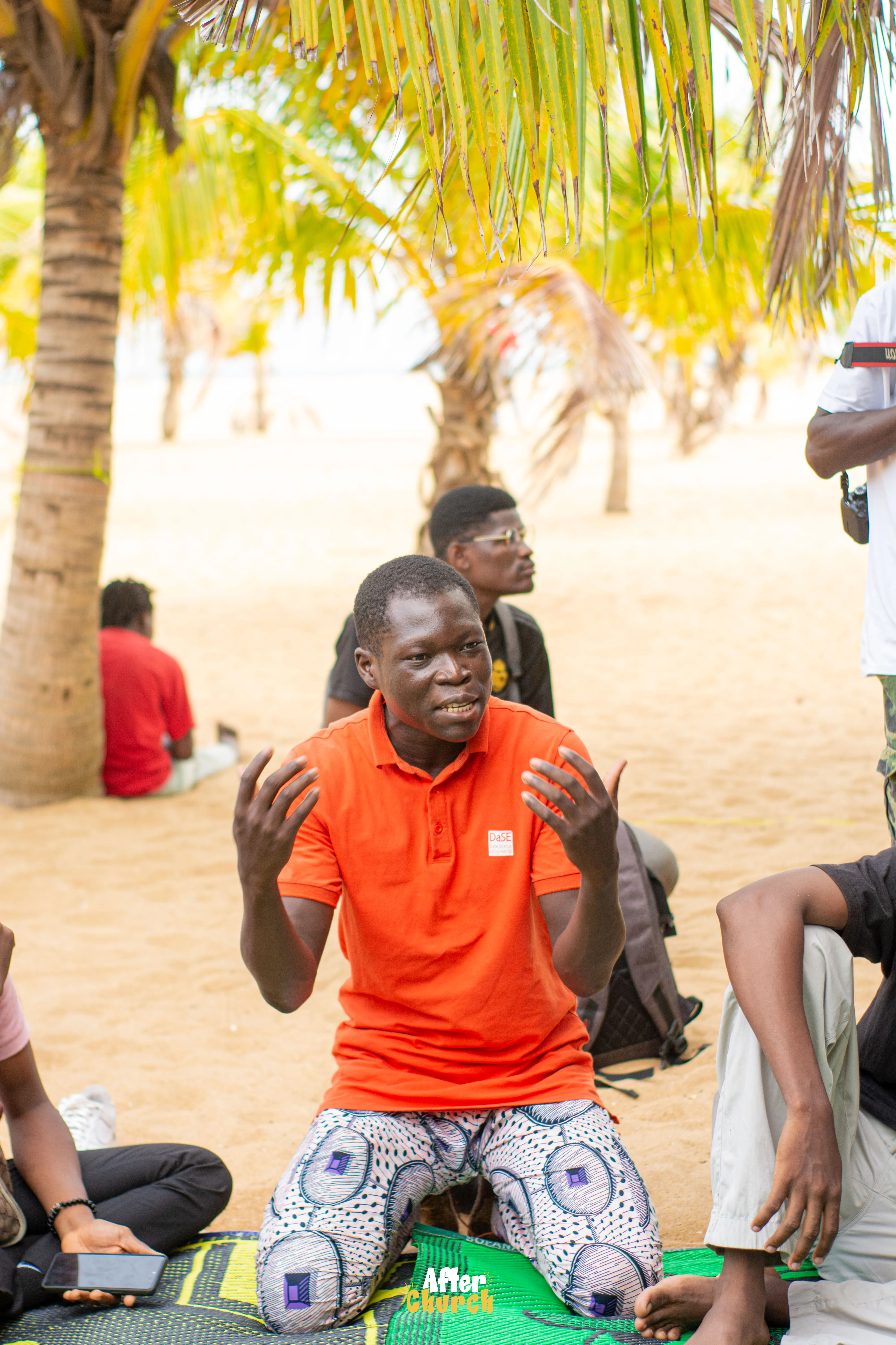 Un jeune homme agenouillé sur une natte, en pleine prise de parole animée devant un groupe assis sous les palmiers, lors d'une session « After Church ».