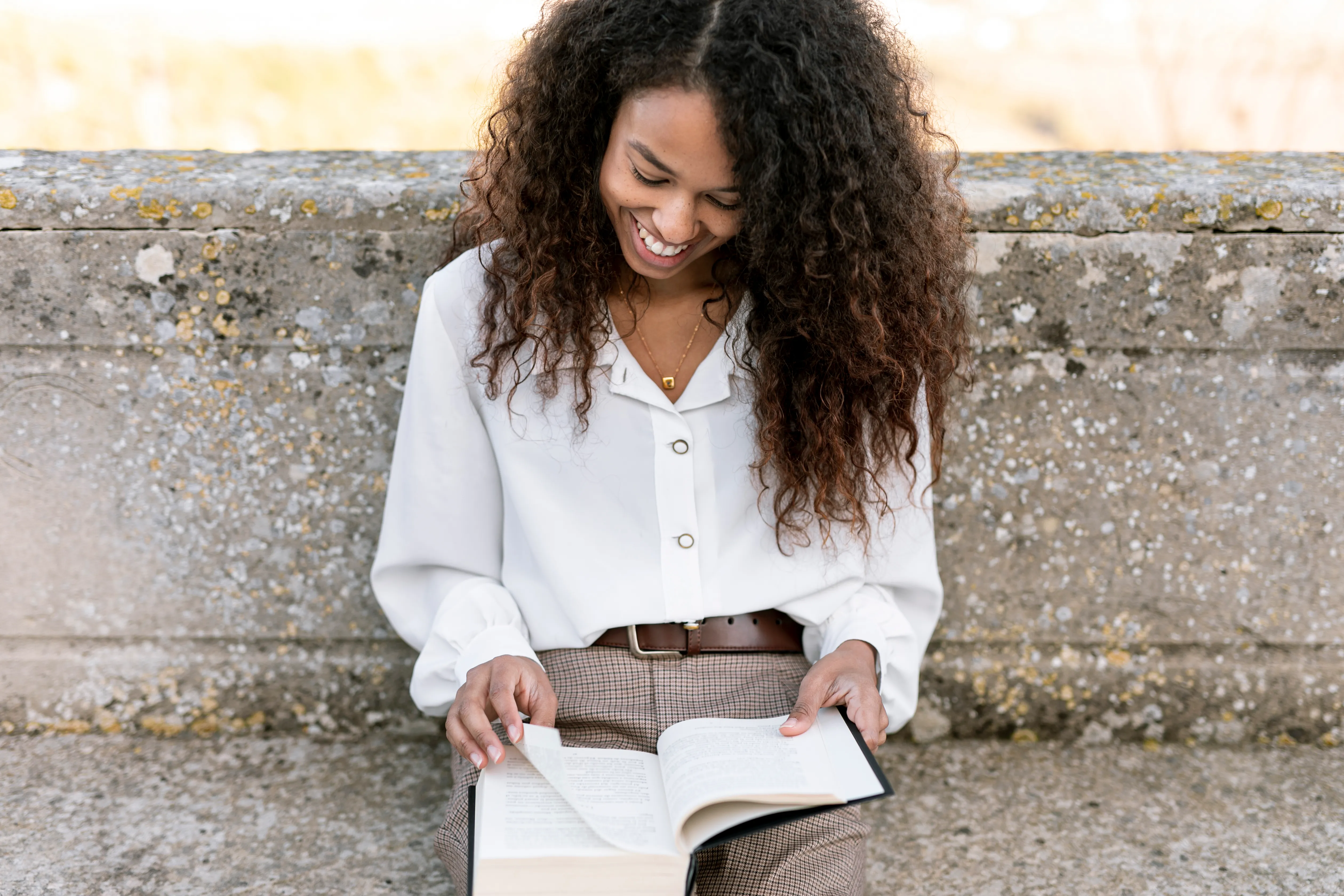 Une jeune femme souriante, lisant un livre à l'extérieur.