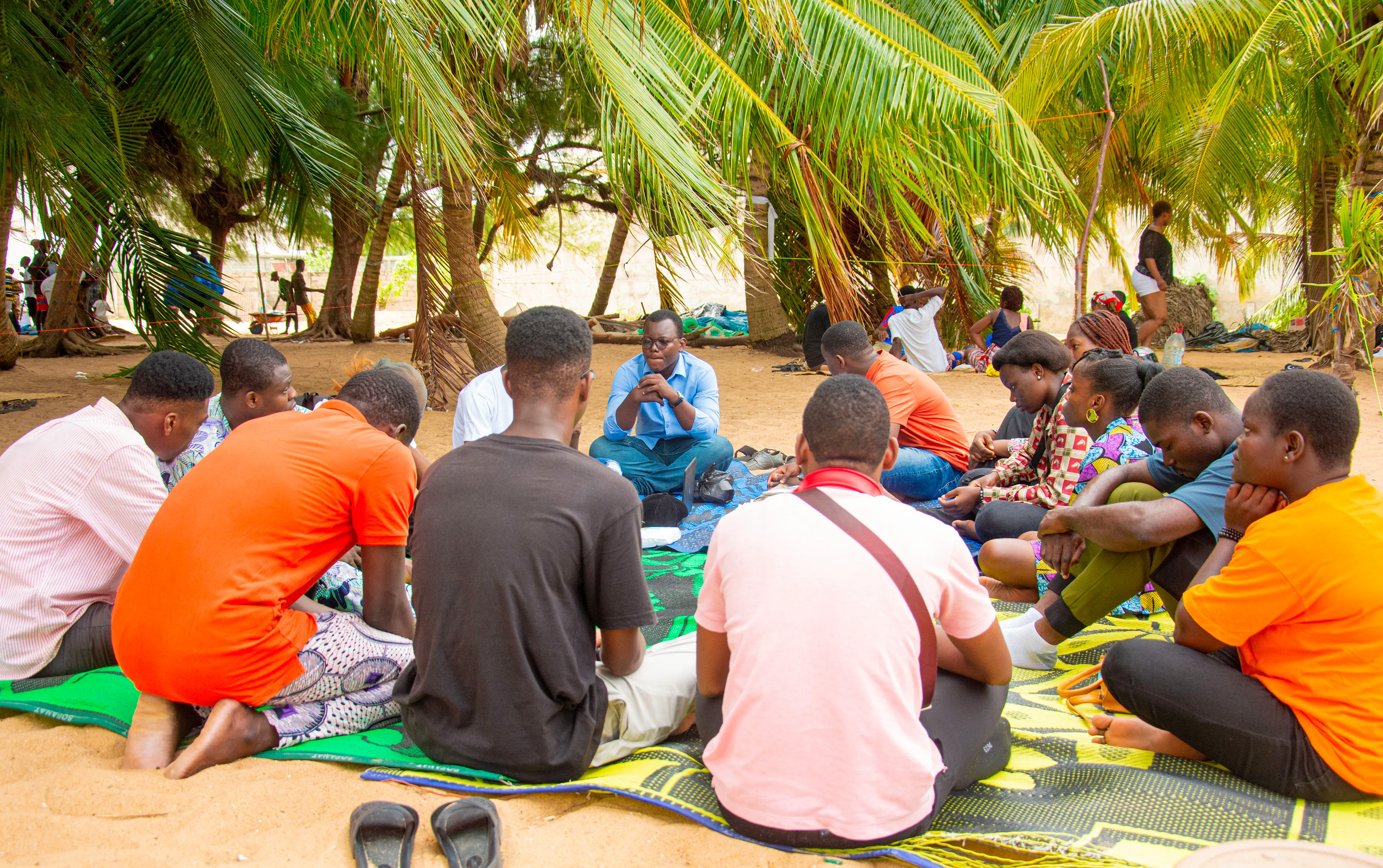 Un groupe de jeunes assis en cercle sur des nattes, sous des palmiers, en pleine discussion.
