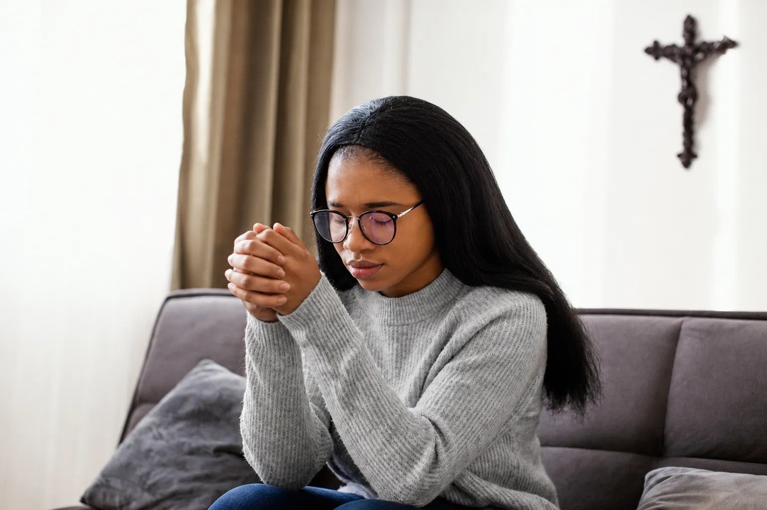Une jeune femme en prière, mains jointes, assise dans son salon près d'une croix murale.