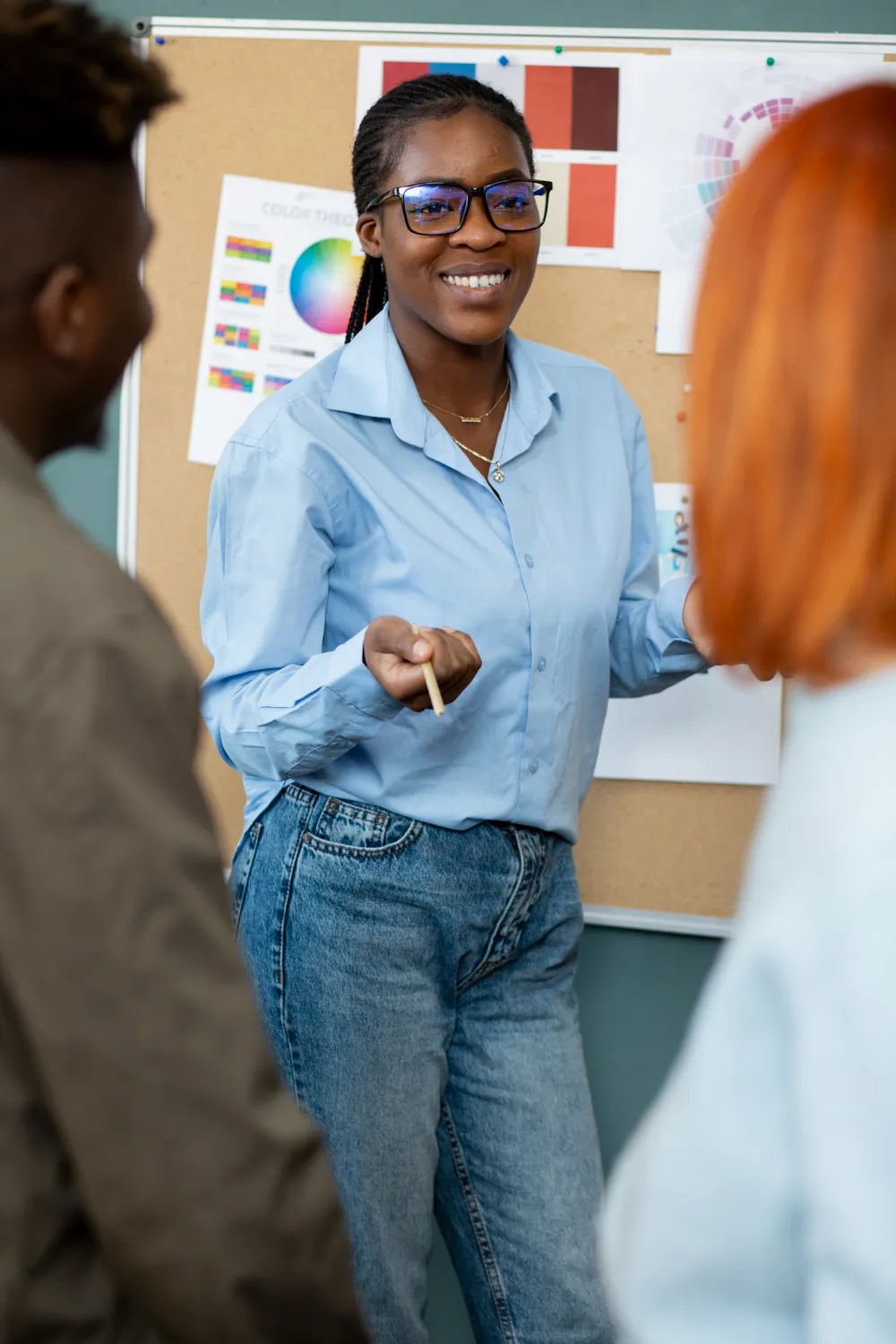 Une jeune femme souriante, en pleine explication devant un tableau de théorie des couleurs, face à deux personnes attentives.