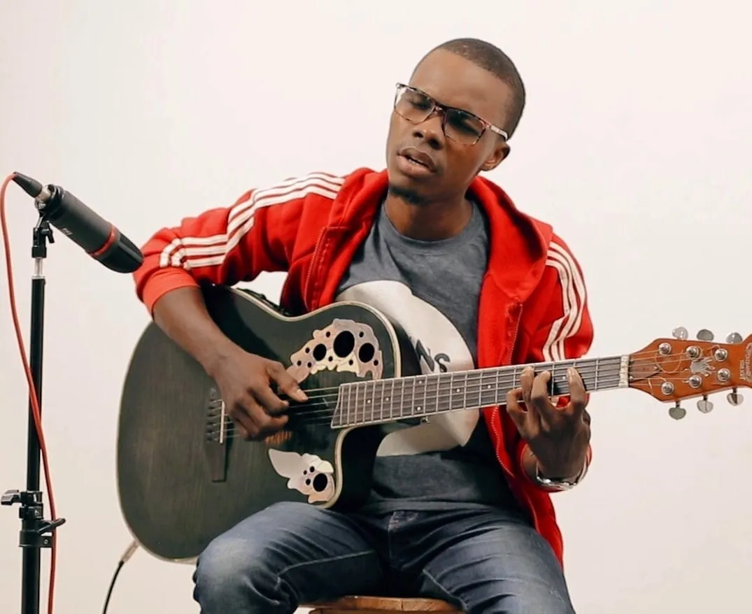 Goussanou Baruc Spinoza en pullover rouge, jouant de la guitare devant un micro.