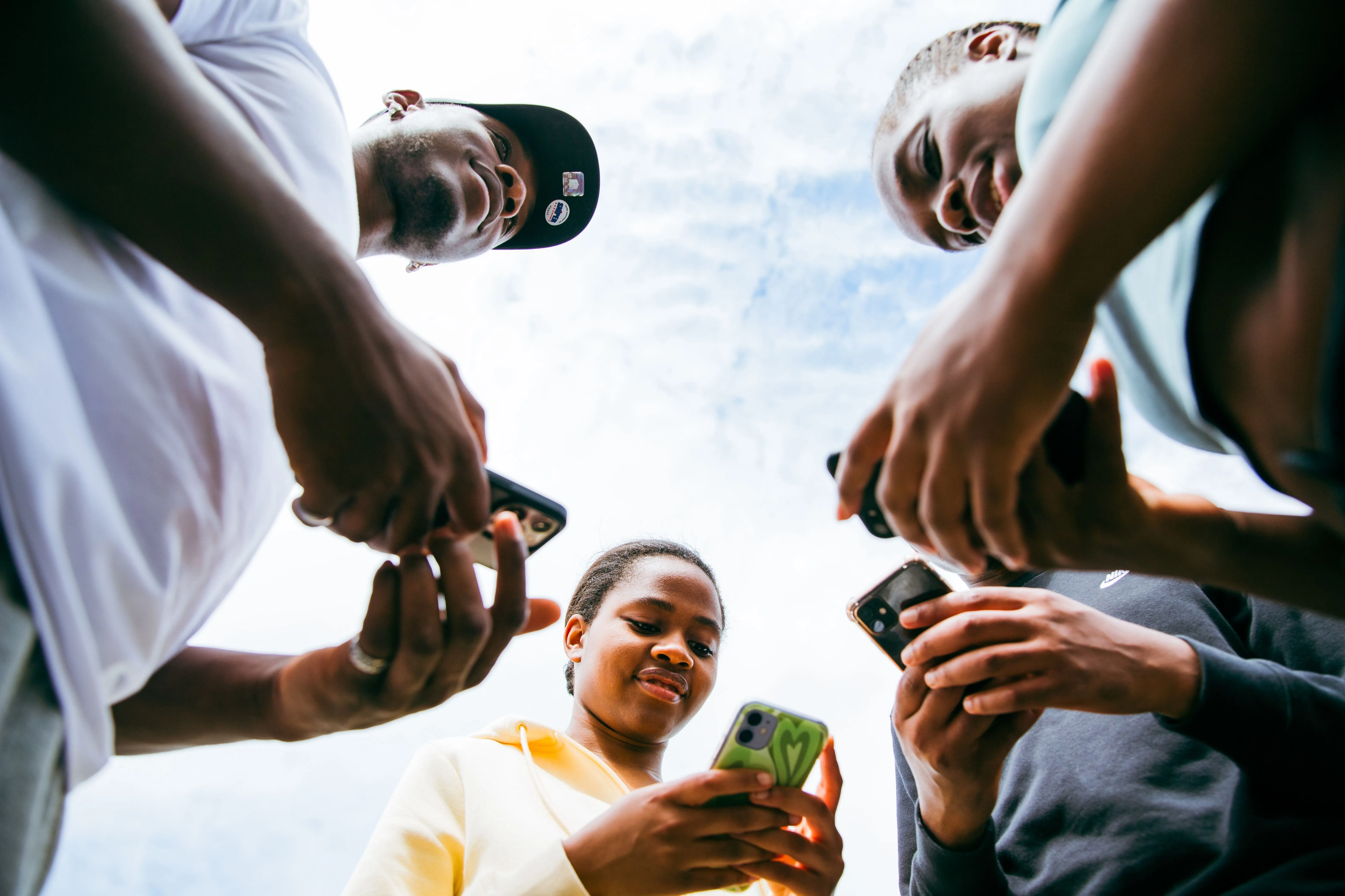 Un groupe de jeunes vu d'en dessous, chacun consultant son smartphone, sous un ciel lumineux.